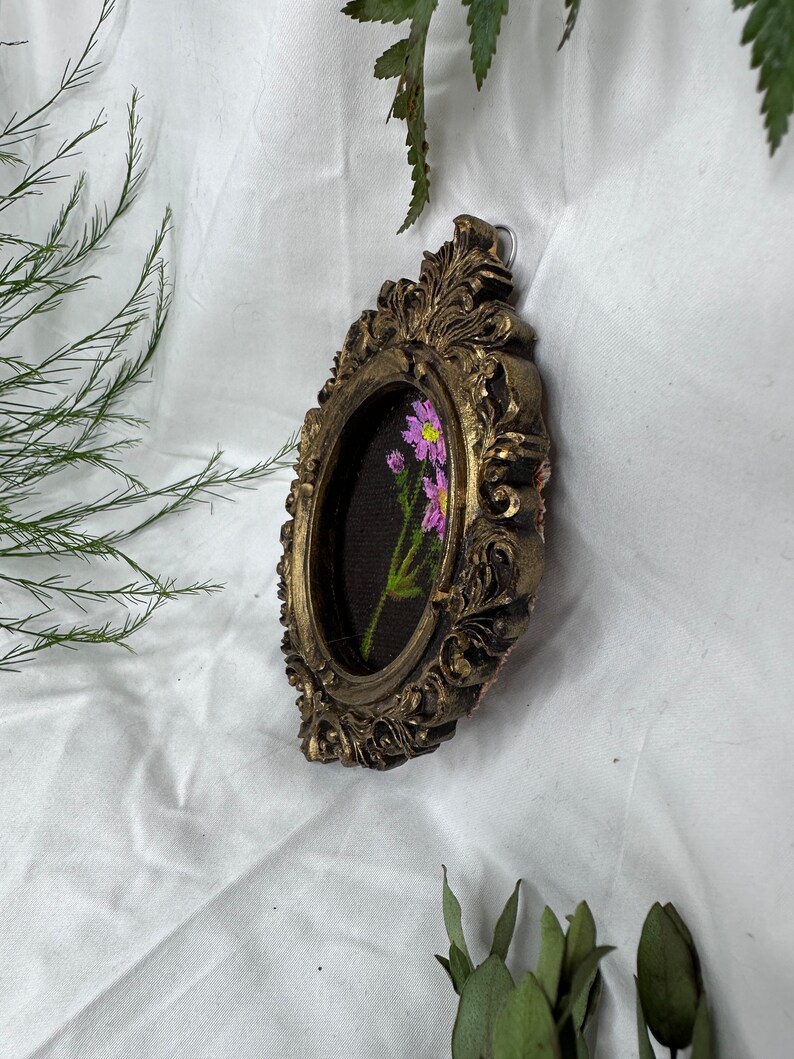 May include: A small oval-shaped picture frame with ornate gold detailing. The frame contains a hand-painted floral design with pink flowers on a dark green background.
