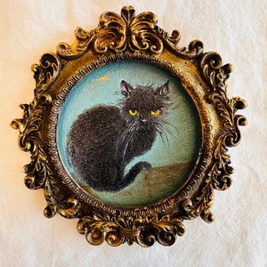 May include: A vintage-style framed painting of a black cat with yellow eyes. The cat is set against a light blue background within an ornate, gold-colored frame. The frame has intricate scrollwork and a detailed design.