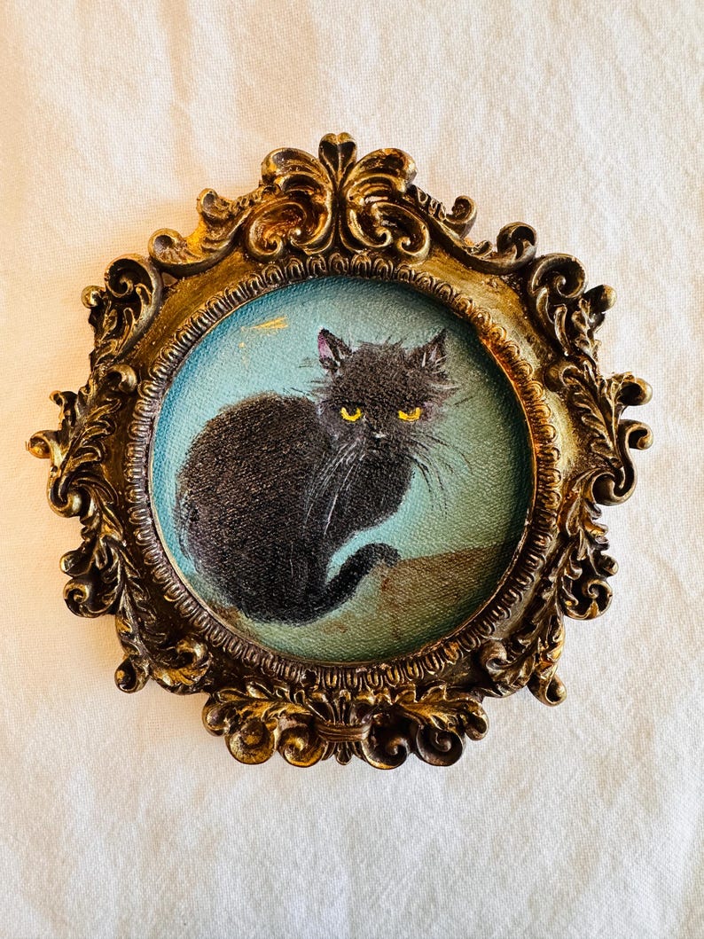 May include: A vintage-style framed painting of a black cat with yellow eyes. The cat is set against a light blue background within an ornate, gold-colored frame. The frame has intricate scrollwork and a detailed design.