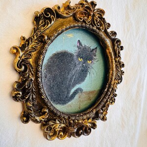 May include: Oval-shaped, antique-style framed painting of a black cat with yellow eyes. The cat is set against a light blue background. The ornate frame is gold-colored with detailed scrollwork.