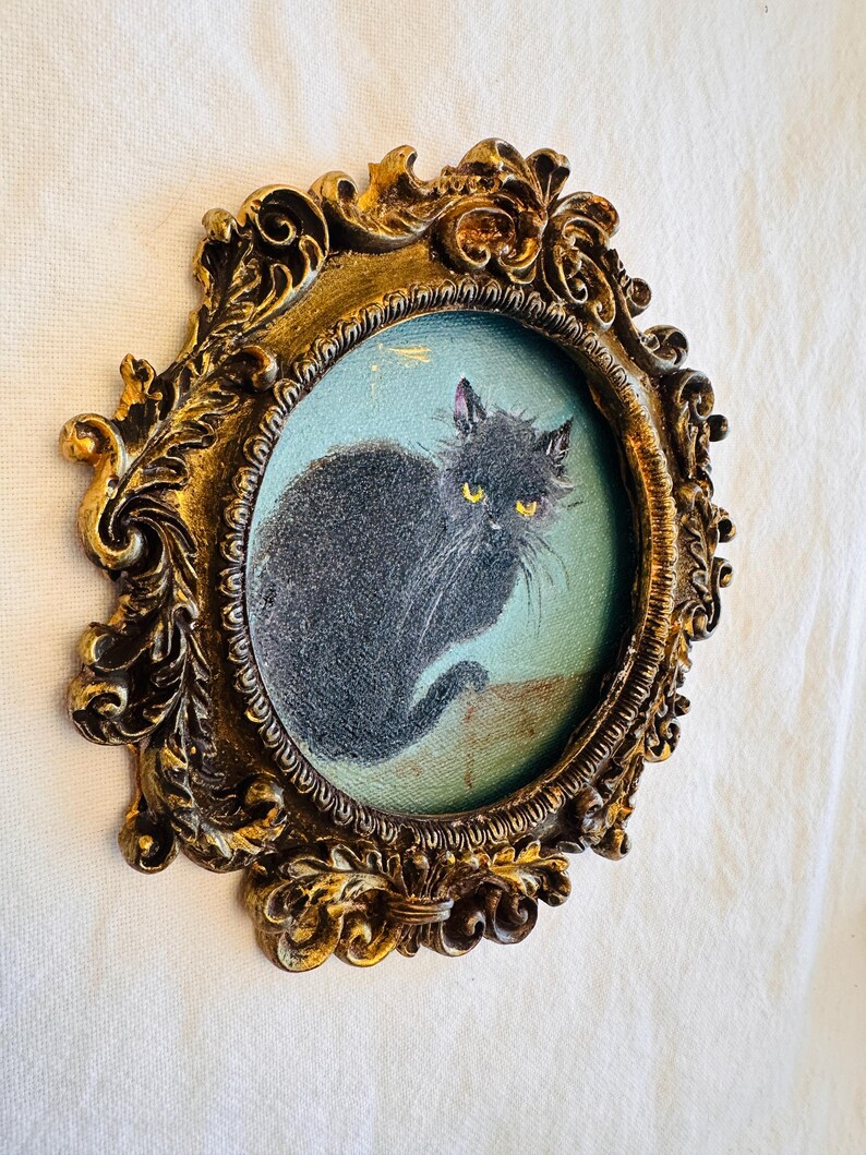 May include: Oval-shaped, antique-style framed painting of a black cat with yellow eyes. The cat is set against a light blue background. The ornate frame is gold-colored with detailed scrollwork.