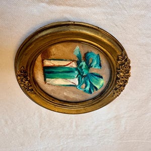 May include: An oval-shaped painting in a gold-toned frame with ornate floral detailing on the sides. The artwork depicts a rectangular white gift box wrapped with a vibrant teal ribbon and a large, prominent bow centered on the front surface.