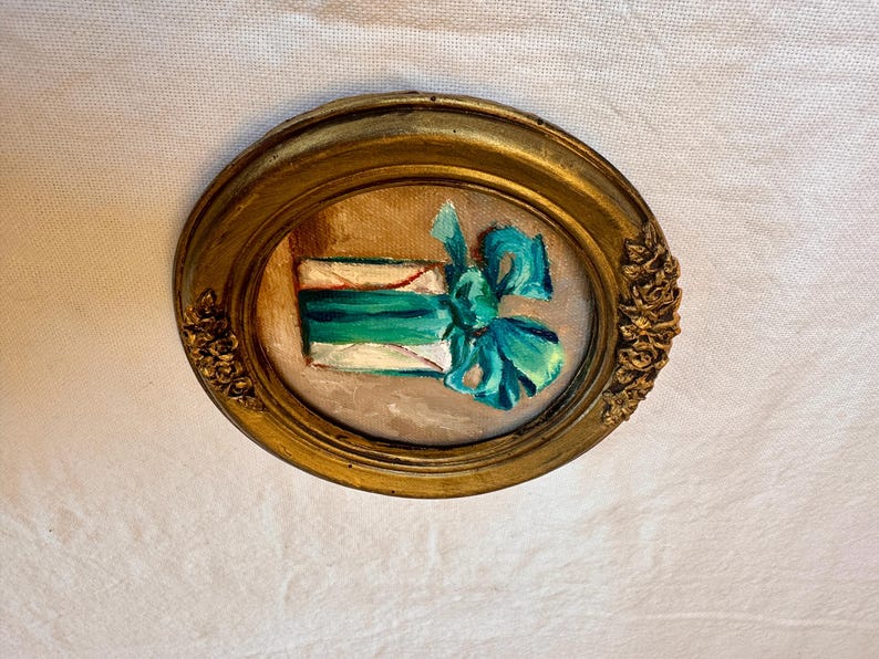 May include: An oval-shaped painting in a gold-toned frame with ornate floral detailing on the sides. The artwork depicts a rectangular white gift box wrapped with a vibrant teal ribbon and a large, prominent bow centered on the front surface.