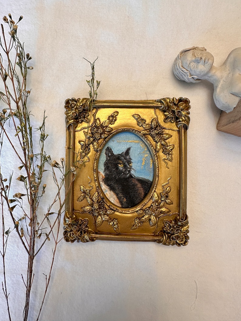K&ouml;nnte beinhalten: Ein gerahmtes Gem&auml;lde einer schwarzen Katze mit gr&uuml;nen Augen vor blauem Hintergrund. Der Rahmen ist verziert und golden mit floralen Details.