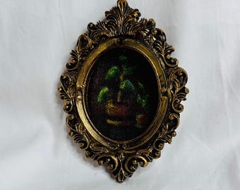 May include: An oval wall hanging featuring a small painting of a green potted plant set within an ornate, metallic gold-colored frame. The frame has intricate scrollwork and decorative relief details surrounding the central dark canvas.