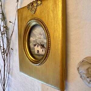May include: A rectangular gold-painted frame with a textured finish and a decorative grape cluster accent at the top. The frame features a central oval opening containing a small oil painting of a white house set against a cloudy, mountainous landscape.