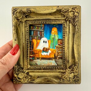 May include: A small, square painting in a gold ornate frame. The artwork depicts a ghost reading a book while seated in an orange armchair. A bookshelf, lamp, and side table are visible in the background. The painting has a whimsical, cozy feel.