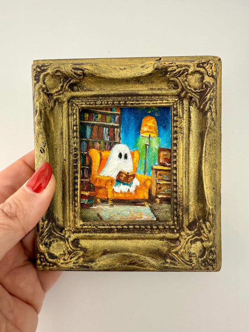 May include: A small, square painting in a gold ornate frame. The artwork depicts a ghost reading a book while seated in an orange armchair. A bookshelf, lamp, and side table are visible in the background. The painting has a whimsical, cozy feel.
