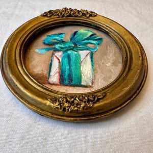 May include: A small circular painting in an ornate gold frame featuring a white gift box wrapped with a vibrant teal ribbon and bow. The artwork is rendered with visible brushstrokes on a textured canvas, set against a neutral beige background.