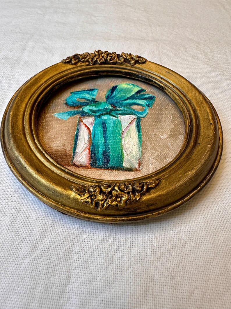 May include: A small circular painting in an ornate gold frame featuring a white gift box wrapped with a vibrant teal ribbon and bow. The artwork is rendered with visible brushstrokes on a textured canvas, set against a neutral beige background.