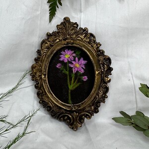 May include: A framed embroidery of a pink flower with green leaves on a black background. The frame is gold and ornate.