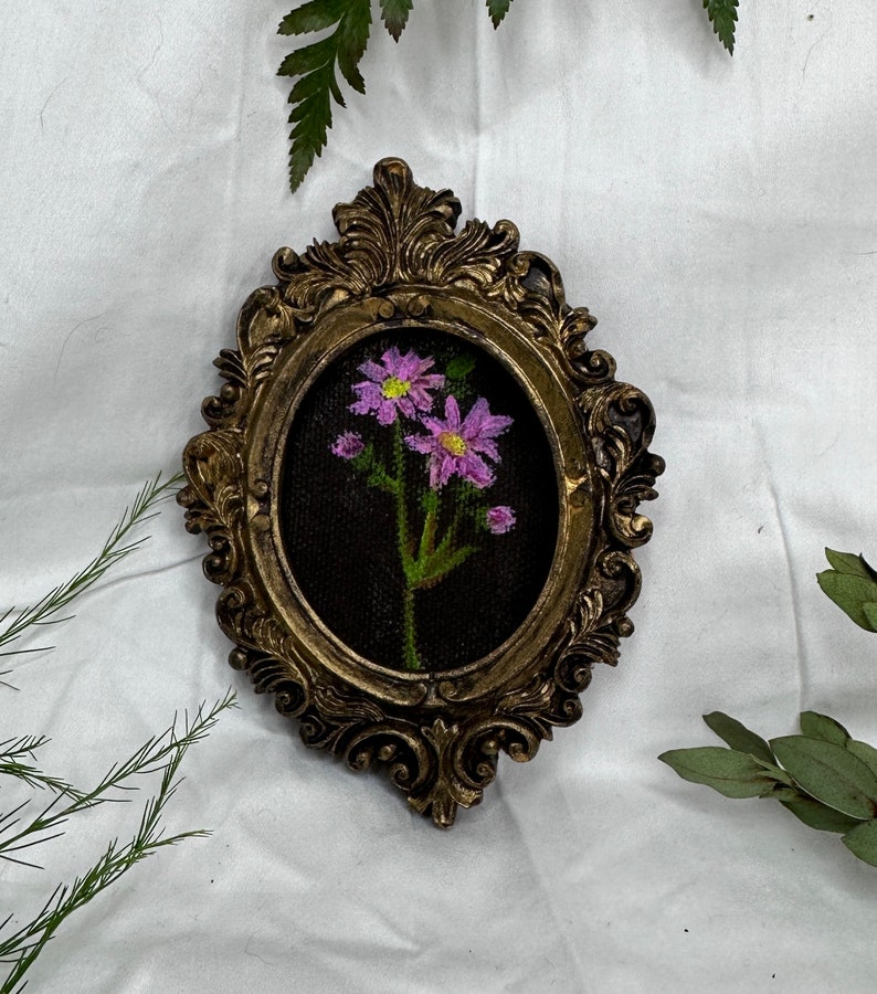 May include: A framed embroidery of a pink flower with green leaves on a black background. The frame is gold and ornate.