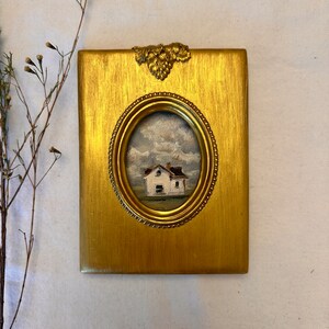 May include: A rectangular gold-toned frame with a metallic grape leaf embellishment at the top center. The frame features an oval opening containing a small oil painting of a white house with a brown roof set against a cloudy sky and green field.