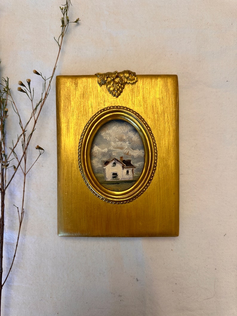 May include: A rectangular gold-toned frame with a metallic grape leaf embellishment at the top center. The frame features an oval opening containing a small oil painting of a white house with a brown roof set against a cloudy sky and green field.