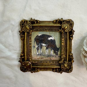 May include: A small square painting of a black and white cow grazing in a field, enclosed in an ornate, antique-style gold frame with intricate scrollwork and bead detailing along the inner border, showcasing a textured, painterly finish.