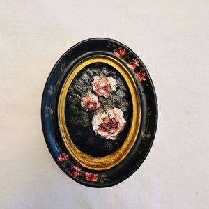 May include: A small oval framed painting of pink roses on a black background. The frame is black with gold trim and has pink roses painted around the edge.