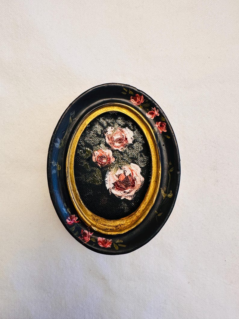 May include: A small oval framed painting of pink roses on a black background. The frame is black with gold trim and has pink roses painted around the edge.