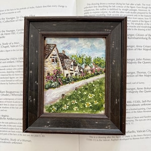 May include: A small oil painting in a dark brown wooden frame depicting a row of stone cottages along a path. The scene features green grass with white flowers, pink bushes, and a light blue sky. The painting is set against a page of text.
