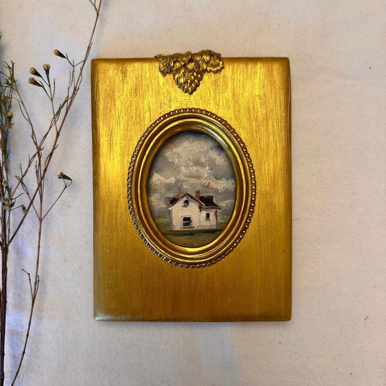 May include: A rectangular gold-toned frame with a textured finish and a decorative grape cluster at the top. The frame features a central oval opening containing a small oil painting of a white house with a brown roof under a cloudy sky.