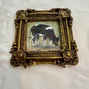 May include: A small square painting of a black and white cow grazing in a field, encased in an ornate, antique-style gold frame with intricate scrollwork and a beaded inner border. The artwork depicts the cow in profile facing to the right.