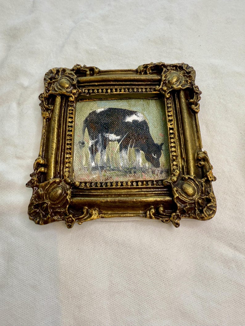 May include: A small square painting of a black and white cow grazing in a field, encased in an ornate, antique-style gold frame with intricate scrollwork and a beaded inner border. The artwork depicts the cow in profile facing to the right.