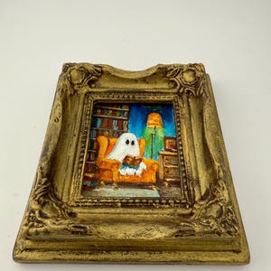 May include: A framed painting depicts a ghost reading in an orange armchair. The artwork is set in a room with a bookshelf and a green lamp. The frame is gold-colored with ornate detailing, creating a whimsical, Halloween-themed piece.