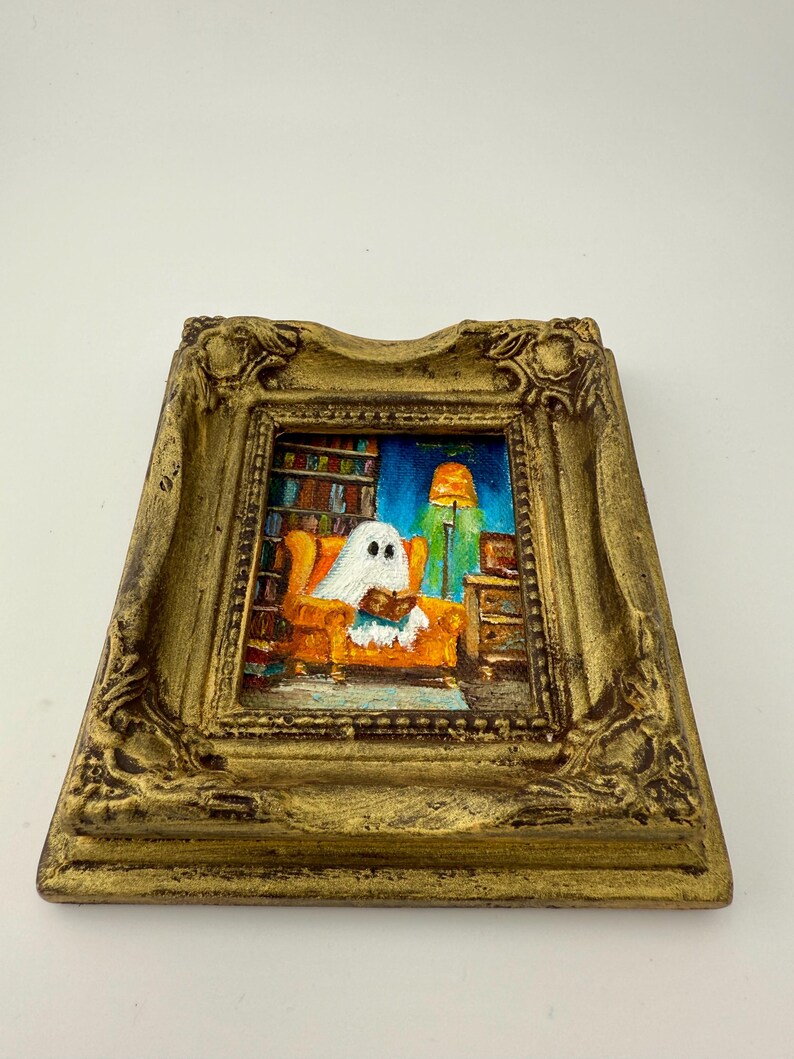May include: A framed painting depicts a ghost reading in an orange armchair. The artwork is set in a room with a bookshelf and a green lamp. The frame is gold-colored with ornate detailing, creating a whimsical, Halloween-themed piece.