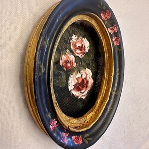May include: Oval framed painting of pink roses on a black background. The frame is black with gold accents and painted with pink roses.
