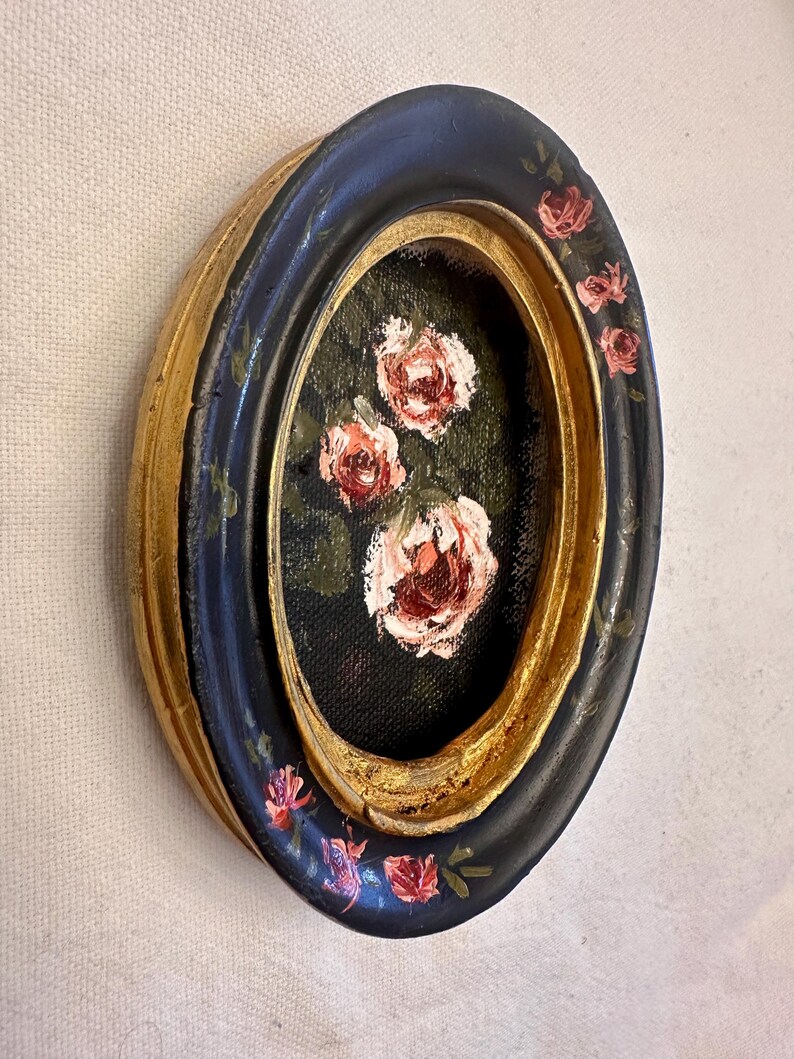 May include: Oval framed painting of pink roses on a black background. The frame is black with gold accents and painted with pink roses.