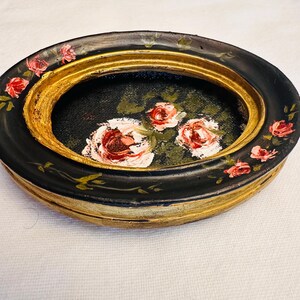 May include: A black oval trinket dish with a gold rim and a painted floral design featuring pink roses on a black background.