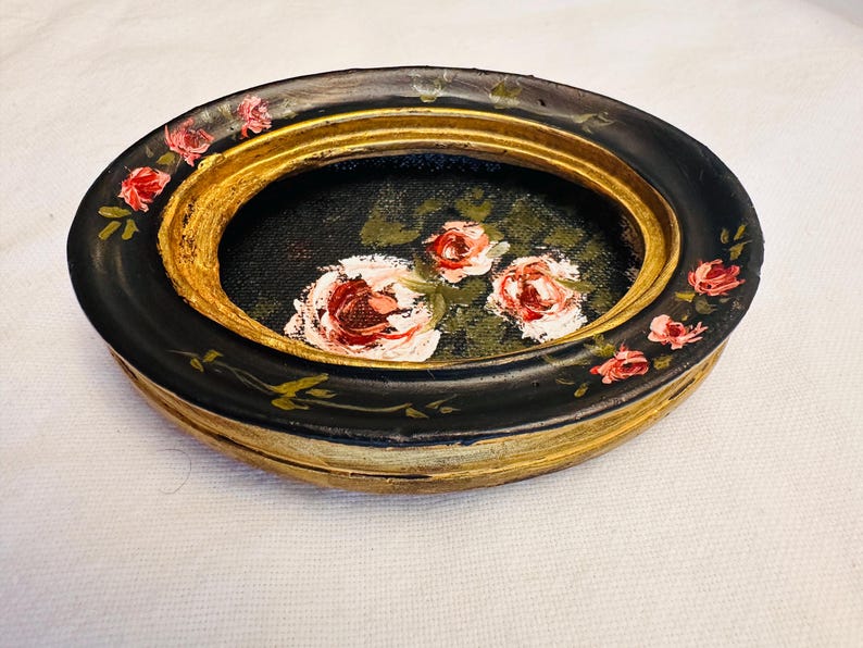 May include: A black oval trinket dish with a gold rim and a painted floral design featuring pink roses on a black background.