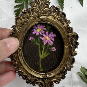 May include: A small, oval-shaped painting of pink flowers with green stems and leaves, framed in a gold ornate frame.