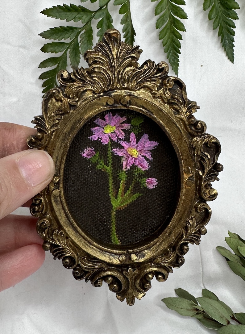 May include: A small, oval-shaped painting of pink flowers with green stems and leaves, framed in a gold ornate frame.