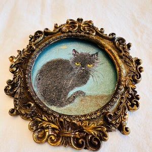 May include: An oval, antique-style frame with a detailed, ornate gold finish. Inside, a small painting depicts a black cat with yellow eyes against a light blue background. The frame has a vintage aesthetic.