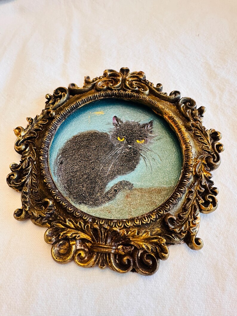May include: An oval, antique-style frame with a detailed, ornate gold finish. Inside, a small painting depicts a black cat with yellow eyes against a light blue background. The frame has a vintage aesthetic.