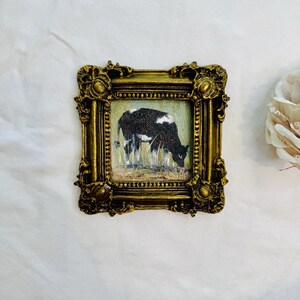 May include: A small square painting of a black and white cow grazing in a field, encased in an ornate, antique-style gold frame with intricate relief detailing and a textured finish. The artwork depicts the cow in profile against a light background.