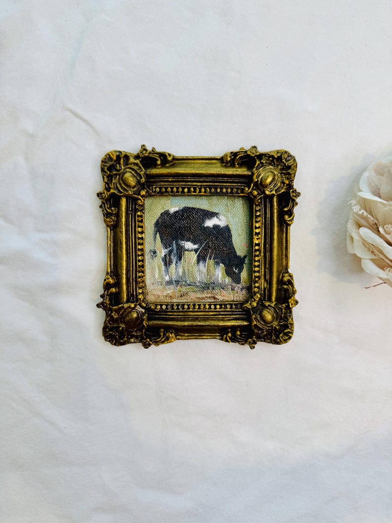 May include: A small square painting of a black and white cow grazing in a field, encased in an ornate, antique-style gold frame with intricate relief detailing and a textured finish. The artwork depicts the cow in profile against a light background.