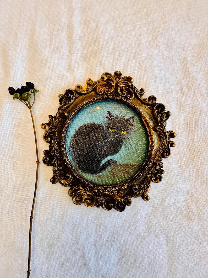 May include: A vintage-style framed print featuring a black cat with yellow eyes. The oval print is set in an ornate, gold-colored frame. A dried plant stem with black berries is to the left of the frame.