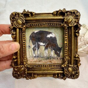 May include: A small square oil painting of a black and white cow grazing in a field, encased in an ornate, heavy gold-toned frame with intricate scrollwork and decorative corner details, featuring a textured canvas surface and visible brushstrokes.