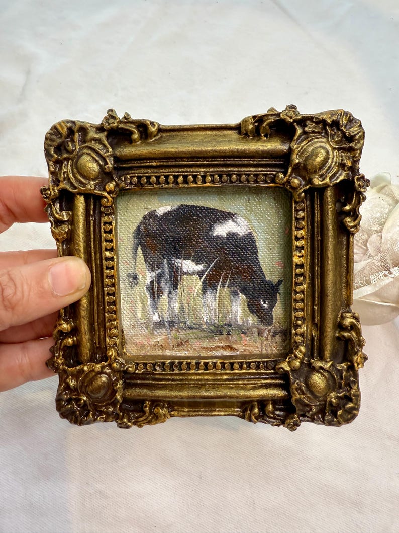 May include: A small square oil painting of a black and white cow grazing in a field, encased in an ornate, heavy gold-toned frame with intricate scrollwork and decorative corner details, featuring a textured canvas surface and visible brushstrokes.