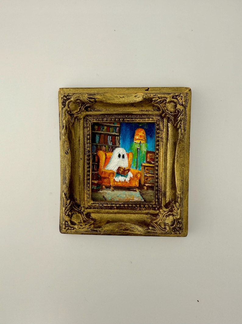 May include: A small framed painting shows a ghost reading in a living room. The ghost is in an orange armchair, with a bookshelf and a lit lamp behind. The frame is ornate and gold-coloured, adding a touch of elegance to the artwork. The painting is a whimsical piece.