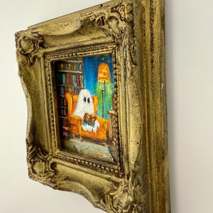 May include: A small, framed painting with a gold ornate frame. The painting depicts a ghost reading in an orange armchair in a room with a bookshelf and a lit lamp. The background is blue.