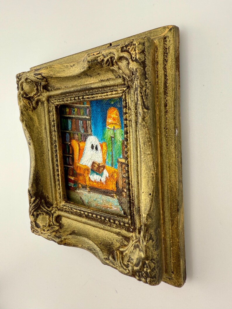 May include: A small, framed painting with a gold ornate frame. The painting depicts a ghost reading in an orange armchair in a room with a bookshelf and a lit lamp. The background is blue.