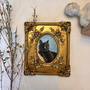 K&ouml;nnte beinhalten: Ein gerahmtes Gem&auml;lde einer schwarzen Katze mit gr&uuml;nen Augen. Der Rahmen ist golden mit verzierten Details und einem floralen Muster. Das Gem&auml;lde ist vor einem hellblauen Hintergrund platziert.