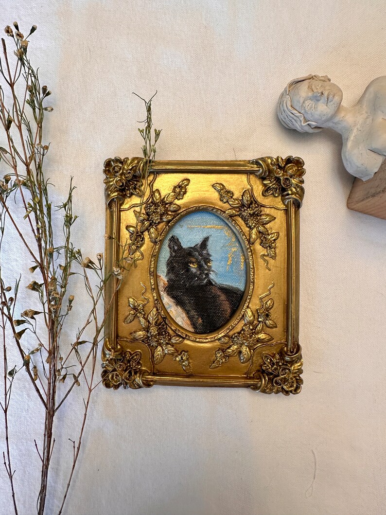 K&ouml;nnte beinhalten: Ein gerahmtes Gem&auml;lde einer schwarzen Katze mit gr&uuml;nen Augen. Der Rahmen ist golden mit verzierten Details und einem floralen Muster. Das Gem&auml;lde ist vor einem hellblauen Hintergrund platziert.