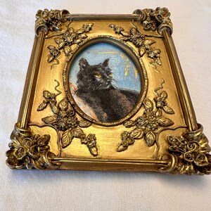 K&ouml;nnte beinhalten: Ein Portr&auml;t einer schwarzen Katze in einem verzierten goldenen Rahmen mit floralen Details. Die Katze schaut nach rechts, ein gelbes Auge ist sichtbar.