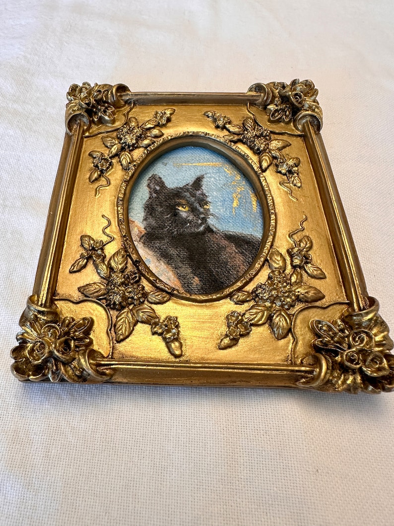 K&ouml;nnte beinhalten: Ein Portr&auml;t einer schwarzen Katze in einem verzierten goldenen Rahmen mit floralen Details. Die Katze schaut nach rechts, ein gelbes Auge ist sichtbar.