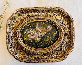 May include: An oval painting of a fawn resting in a field of wildflowers, set within a gold-toned, ornate, rectangular frame. The painting features a fawn with white spots, lying in green grass with yellow and pink flowers.