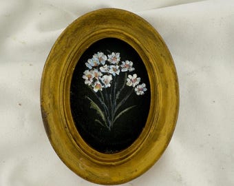 May include: An oval-shaped, mustard-yellow wooden frame containing a small painting of a bouquet of white flowers with yellow centres against a solid black background. The frame has a textured, weathered finish with visible brush strokes throughout.