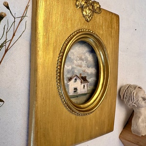 May include: A rectangular gold-painted frame with a central oval opening featuring a beaded inner edge and a decorative leaf motif at the top. The frame contains a small oil painting of a white house under a cloudy sky with visible brushstrokes.